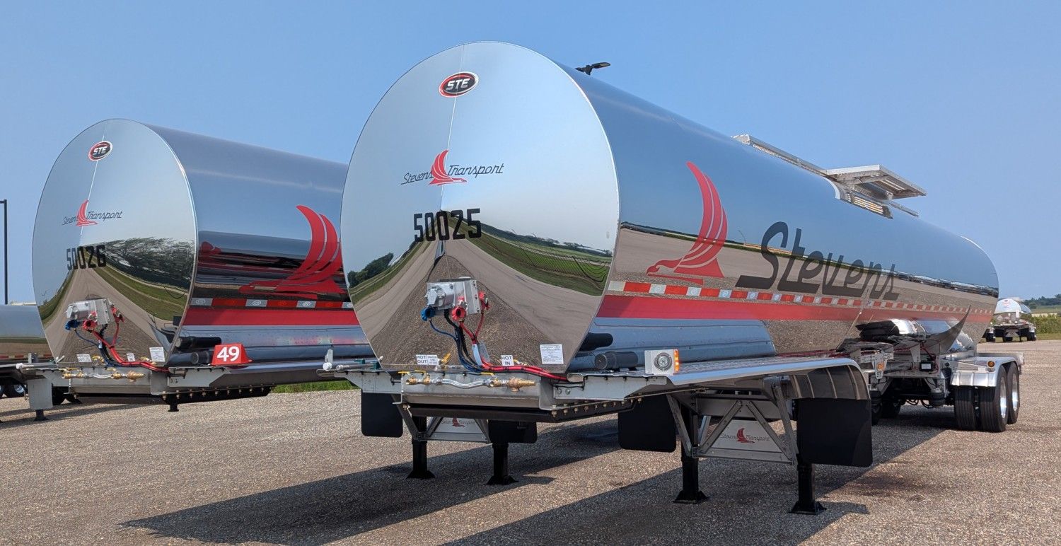Two polished stainless steel Stevens Transport tanker trailers parked side by side reflecting the surrounding landscape