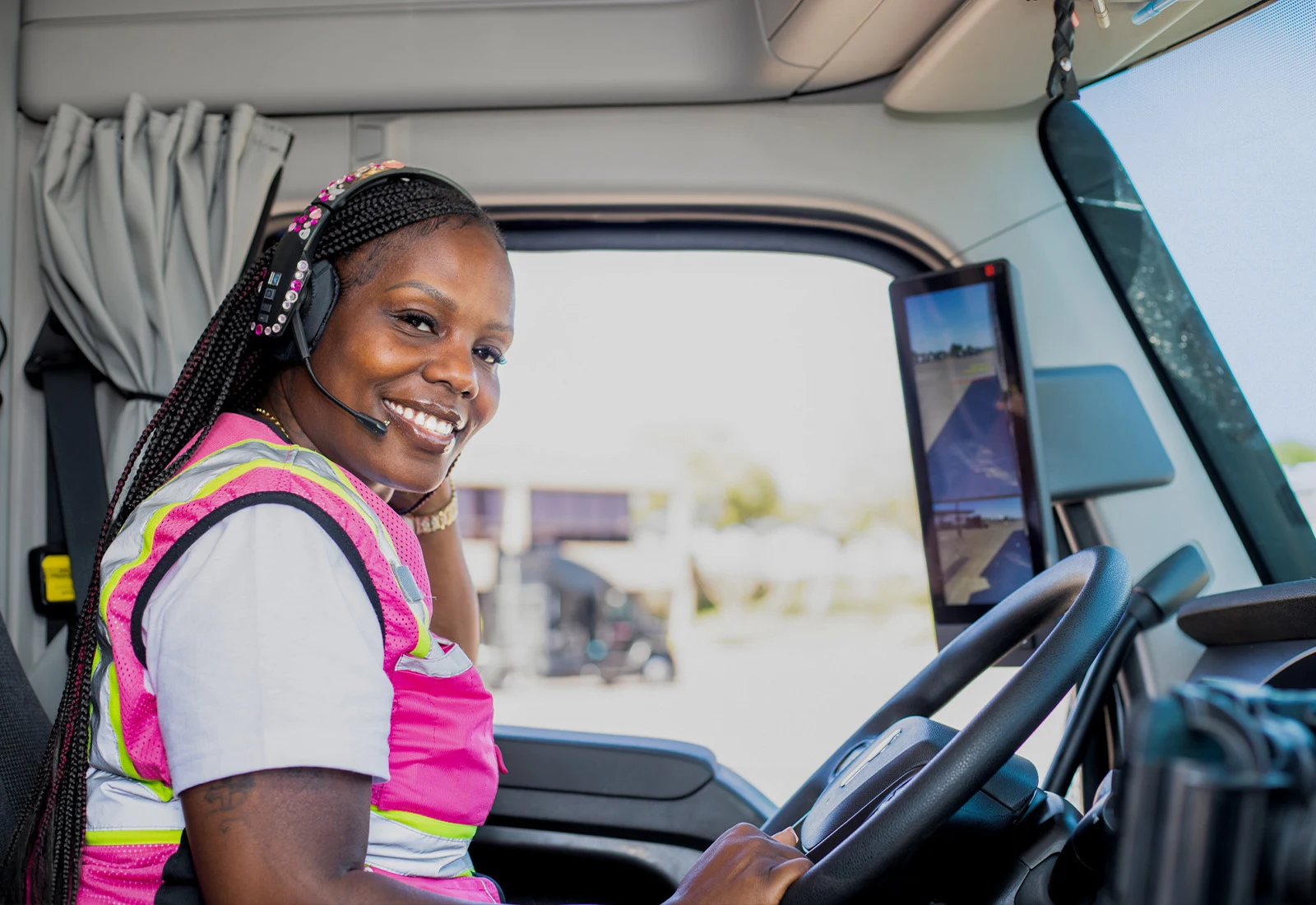 Female trucker with pink safety vest and headset smiling in semi truck drivers seat