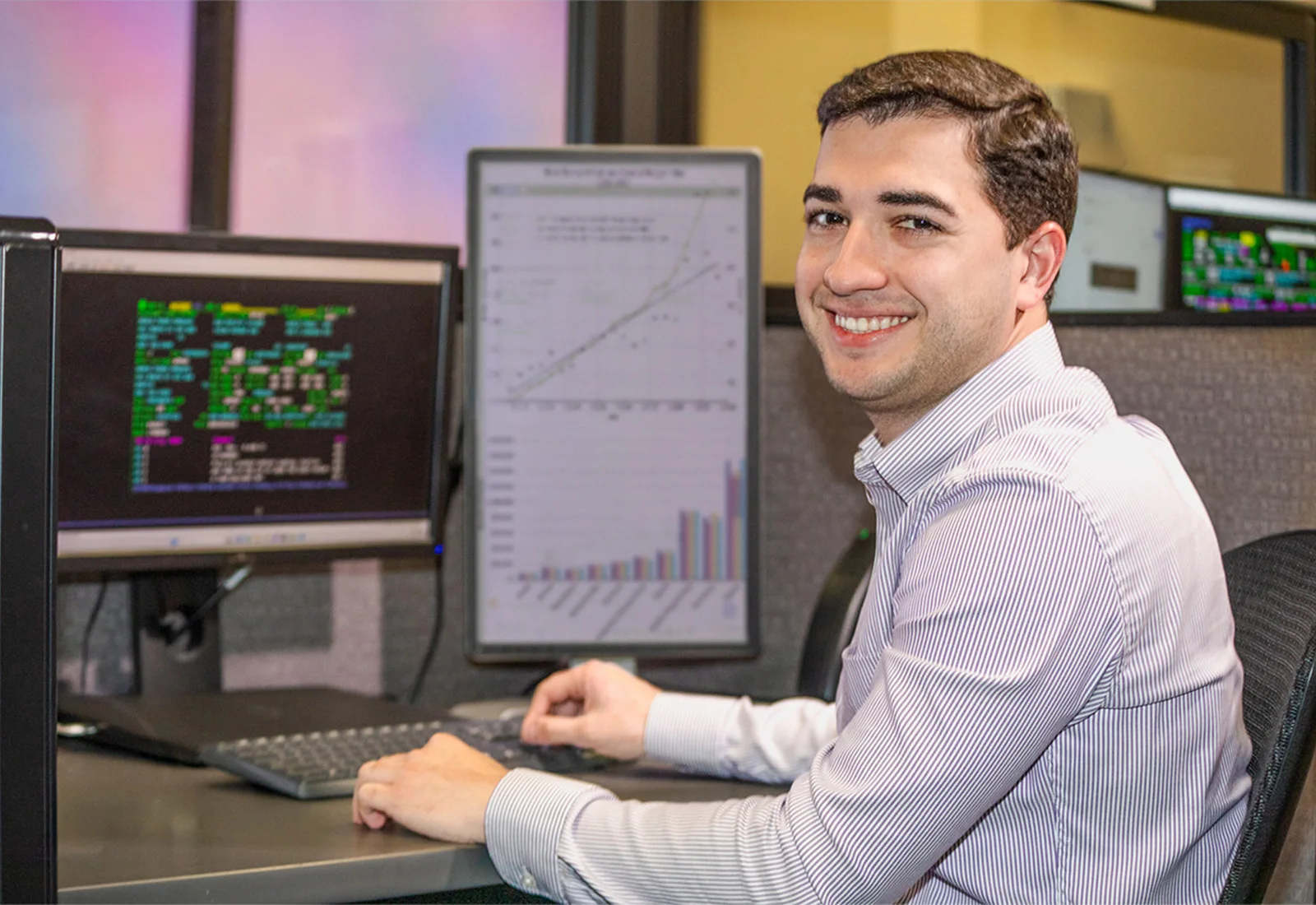 A smiling logistics professional seated at a desk with multiple monitors displaying data, charts, and graphs in an office.