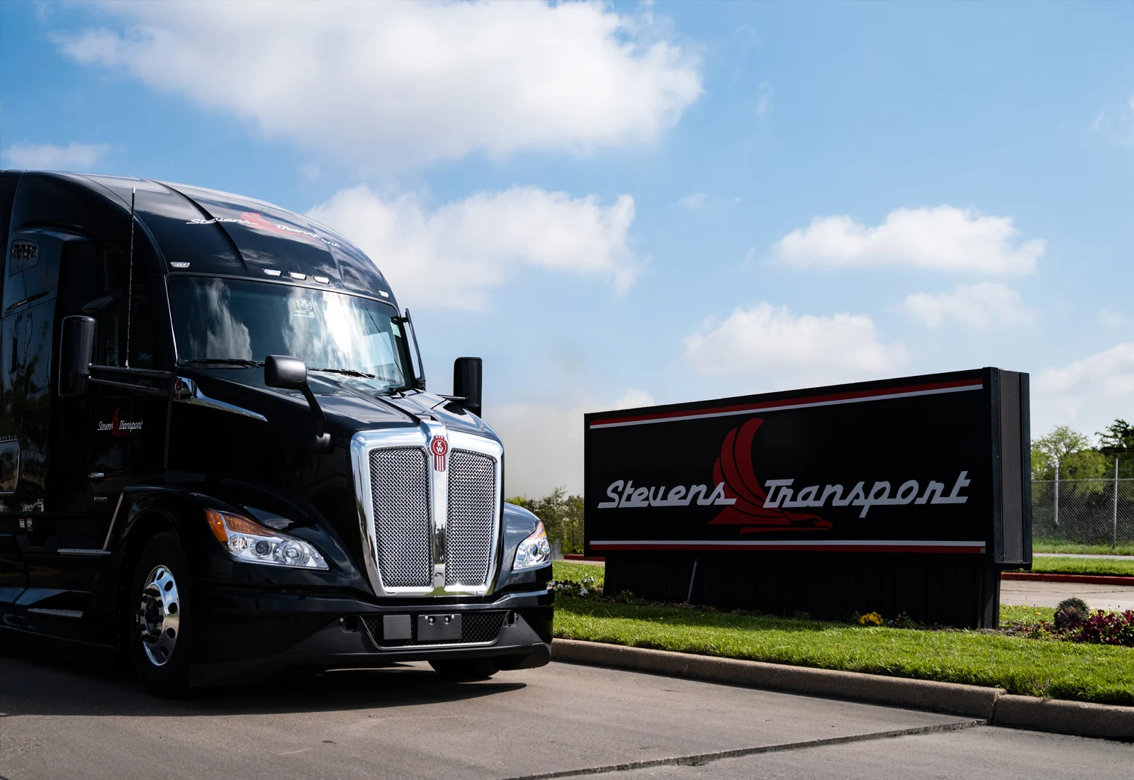 A black Stevens Transport Kenworth semi truck parked beside the Stevens Transport entrance sign on a landscaped lawn under a blue sky with scattered clouds.