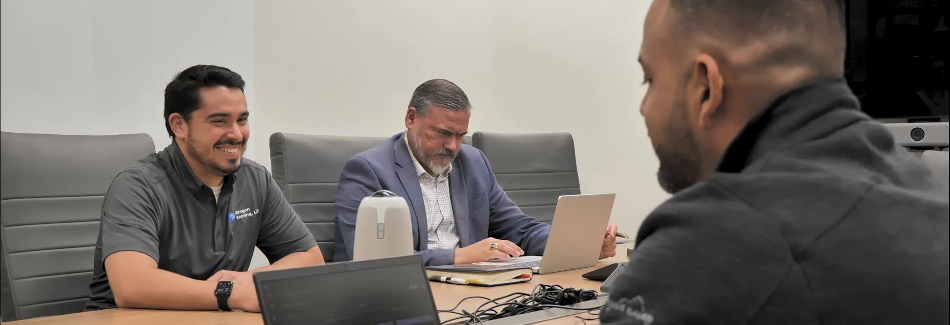 Three professionals seated around a conference table with laptops during a meeting, two of them smiling and one working on a laptop.