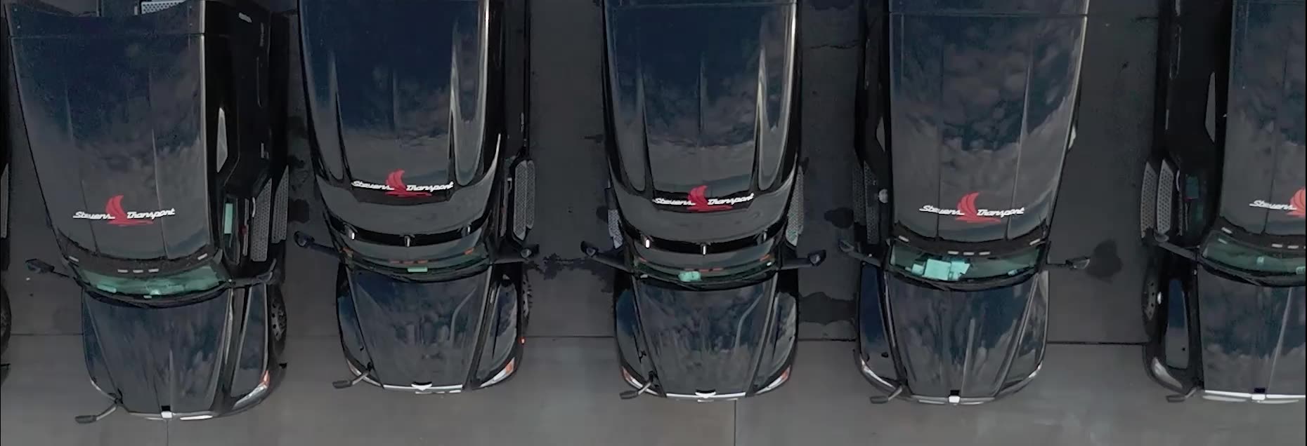 Aerial view of a row of black Stevens Transport semi-truck cabs parked side by side in a lot.