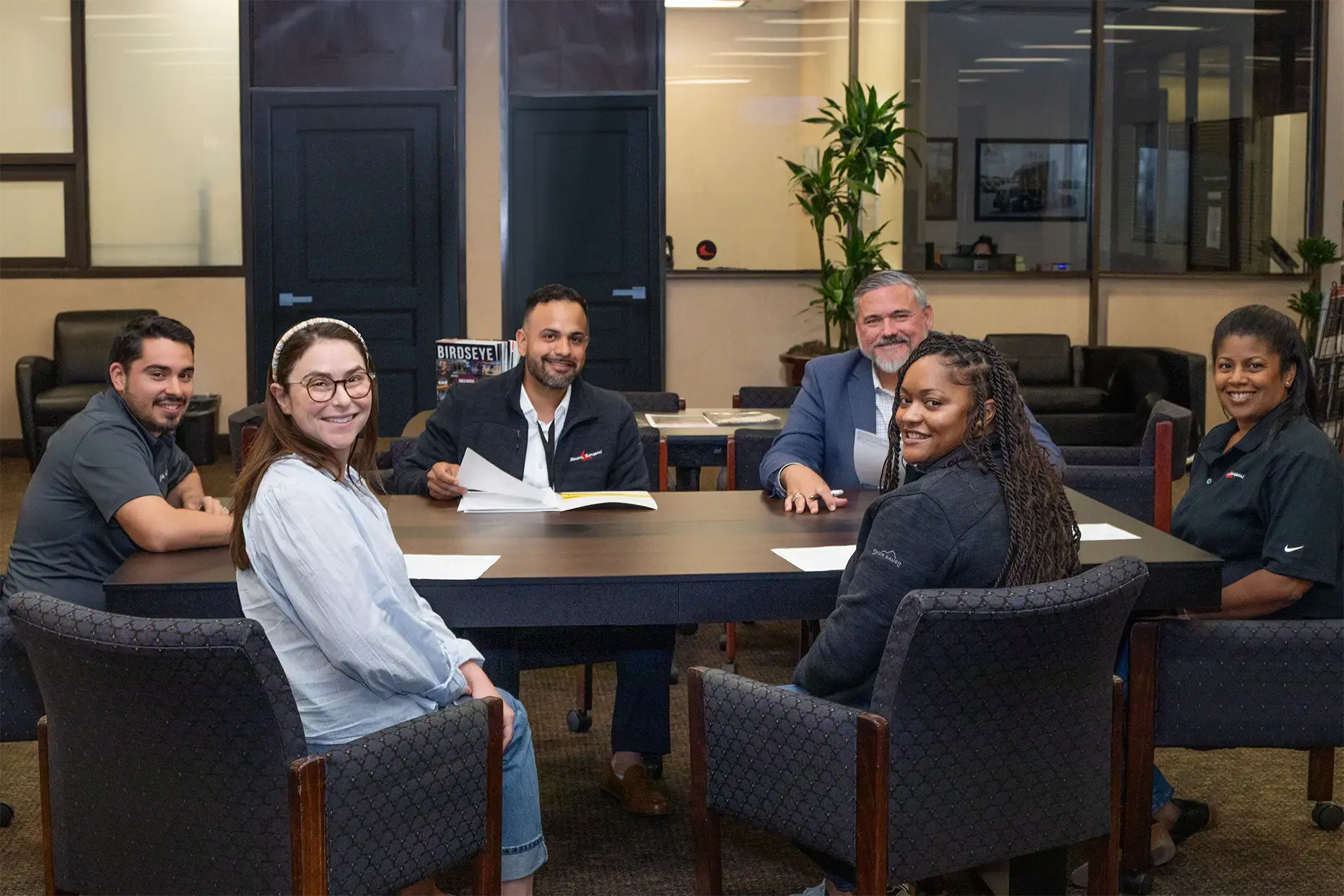 Six smiling corporate employees, several in company-branded polos and jackets, sit around a conference table with papers in a modern office lounge. They turn to face the camera.
