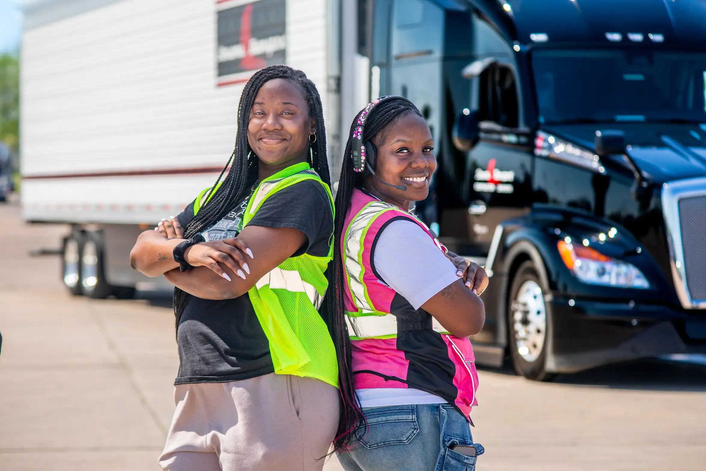 Two smiling women truck drivers stand back to back with their arms crossed, one in a neon yellow safety vest and the other in a pink safety vest and headset, in front of a black semi truck and white trailer.