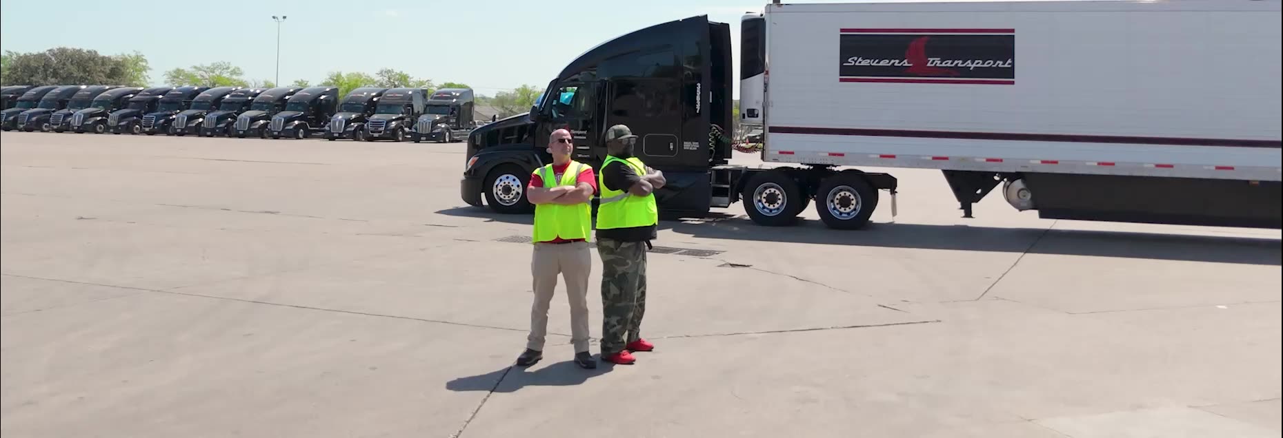 Two Stevens Transport drivers in safety vests standing in a truck yard beside a black semi-truck and white trailer, with a row of parked trucks in the background.