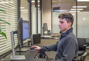 A young intern in a gray fleece pullover works at a dual-monitor standing desk, using a mouse while viewing a spreadsheet, in an office with frosted glass partitions.
