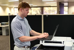 A young intern in a striped polo shirt types on a laptop resting on top of a copier in an office with cubicle partitions and large windows behind him.